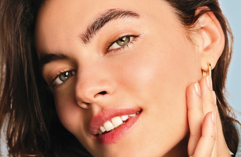 Mulher sorridente de cabelos e olhos castanhos, com a pele hidratada e sem linhas fina por conta do ácido hialurônico.