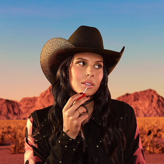 Mulher de cabelos castanhos e longos usa roupa e chapéu pretos com pontos brilhantes e aplica Gloss Labial na boca. Ela usa esmalte escuro, olha para a direita e está em fundo de montanhas com céu azul.