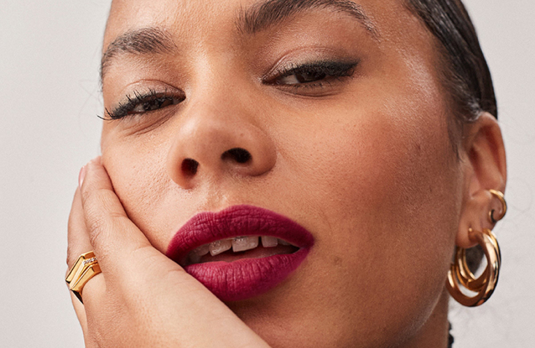 Mulher com os lábios entreabertos com batom na cor vinho e maquiagem para festa com as pálpebras brilhosas, apoia a mão esquerda na bochecha e olha para frente.