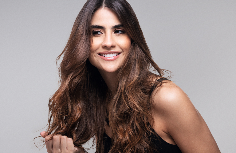 Mulher sorridente com longos cabelos castanhos ondulados após um banho de brilho nos fios, segurando suavemente uma das mechas com a mão direita, usando uma regata preta contra um fundo cinza neutro.