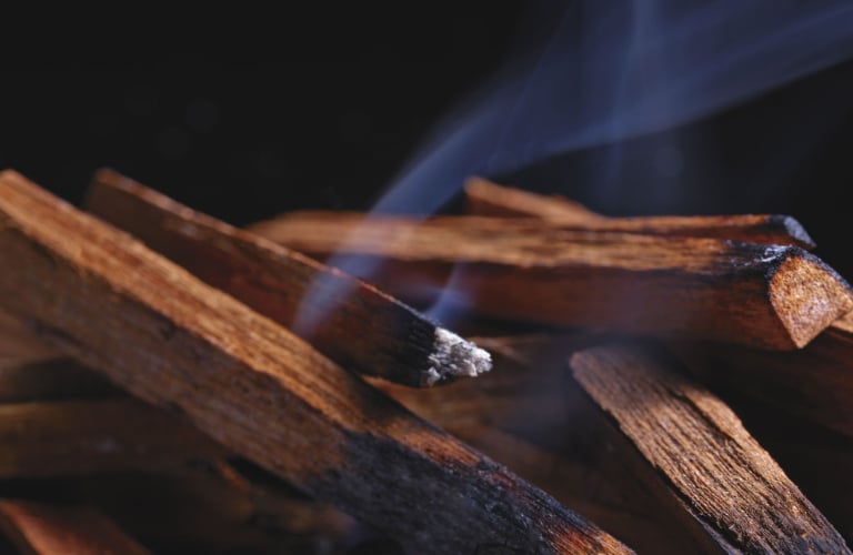Pequenos pedaços de madeira empilhados queimando levemente e soltando fumaça branca contra um fundo escuro, pelo aroma amadeirado em perfumes com feromônio.