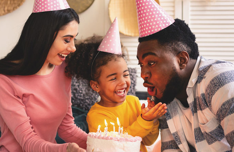 Uma mulher e uma criança com chapéus rosa de aniversário seguram um bolo com velas comemorativas, enquanto um homem, que também usa chapéu rosa, sopra as velas do bolo. As pessoas estão em uma sala de estar com paredes brancas, sofá cinza e decoração monocromática.