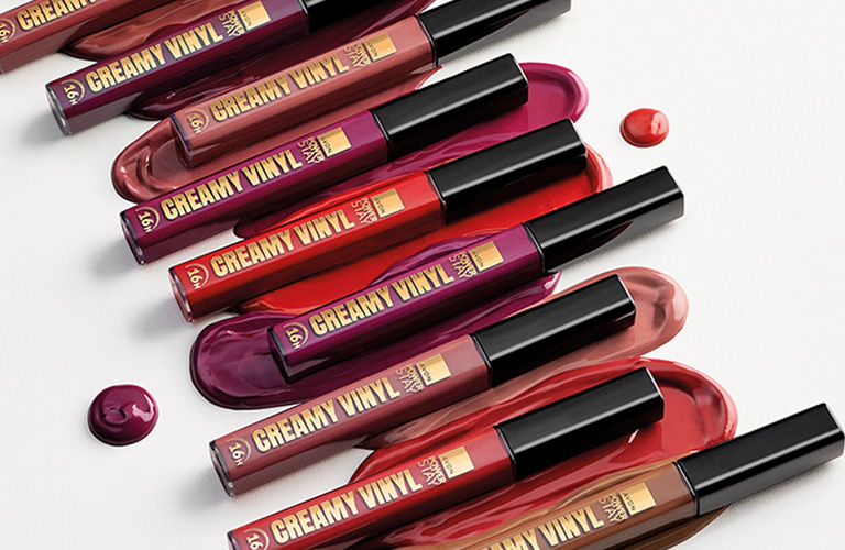 Batons líquidos da Avon Power Stay Creamy Vinyl em variados tons de vermelho, vinho e marrom, dispostos diagonalmente sobre amostras cremosas de suas cores em fundo branco.