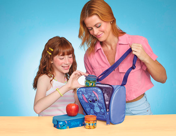 Menina e mulher, ambas de cabelos ruivos, interagem com itens e alimentos em produtos da coleção Stitch. O fundo é azul e a bancada é em tom de madeira.