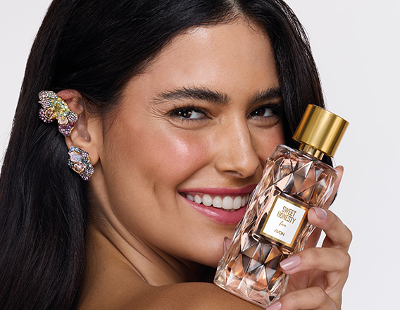 Mulher branca de cabelos castanhos segura frasco da fragrância Sweet Honesty Fun com tampa dourada e líquido translúcido. Ela sorri e está em fundo claro.