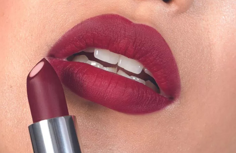 Close-up dos lábios de uma pessoa com batom matte em tom de vinho. Ela segura o bastão do batom à frente, destacando a cor intensa e o acabamento aveludado.
