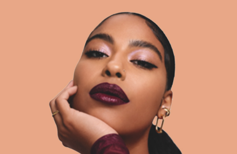 Mulher de cabelos curtos escuros vestindo colete rosa, com maquiagem suave para o trabalho e delineado gatinho, posando com a cabeça inclinada em um fundo branco.