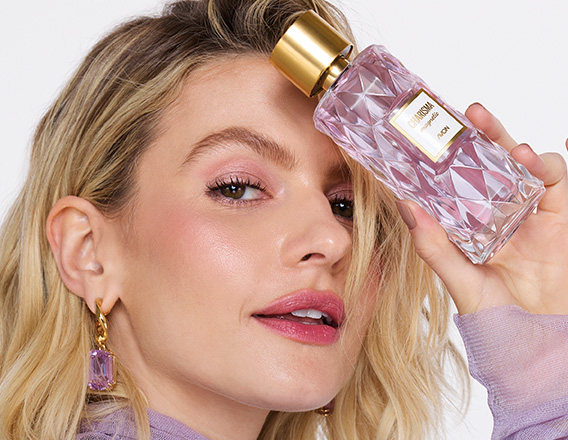 Mulher loira segura frasco da fragrância Charisma Magnetic com tampa dourada e líquido lilás. Ela usa lilás e está em fundo claro.