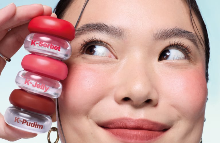 Mulher de descendência asiática sorrindo. Ela possui bochechas levemente rosadas, pele viçosa e lábios pintados com um batom de tom avermelhado. A modelo olha para o alto e segura, próximo ao rosto, uma pilha vertical de três pequenos potes transparentes de lip tint da marca Avon Sweets.