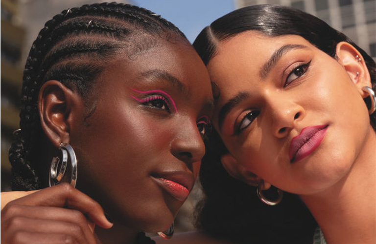 Duas mulheres jovens abraçadas usam maquiagem natural e leve. A primeira mulher tem pele negra, usa tranças e jaqueta esportiva colorida, e a outra mulher tem pele clara, cabelos castanhos e veste blusa verde.