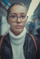 Foto de mulher branca de cabelos presos, com blusa de gola alta branca e jaqueta de couro preta olhando para a câmera. O fundo é a imagem desfocada do interior de um vagão de metrô.
