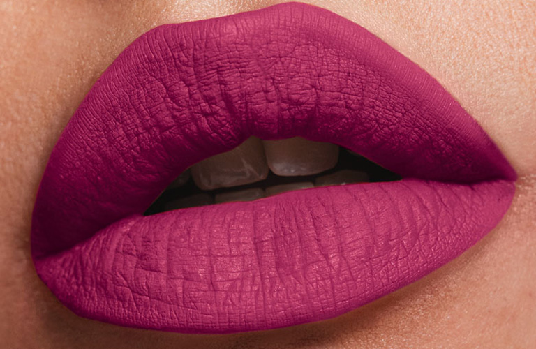 Close-up nos lábios de uma pessoa usando batom matte magenta intenso. A pele é clara e os lábios estão levemente entreabertos, destacando a textura aveludada do produto.