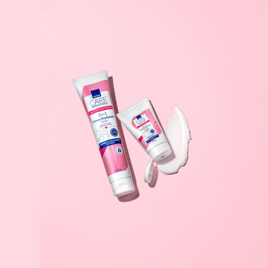 Bisnagas branca com rosa de Cremes Depilatórios; uma grande fechada à esquerda e outra pequena, à direita, com a tampa aberta e textura branca do produto. Fundo rosa liso.