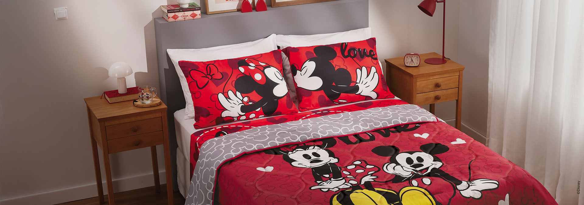 Banner em fundo claro, uma cama com roupa de cama e travesseiros estampados com Mickey e Minnie, ao lado de uma mesa de cabeceira. Texto: "Produtos que trazem aconchego e elegância para sua casa."