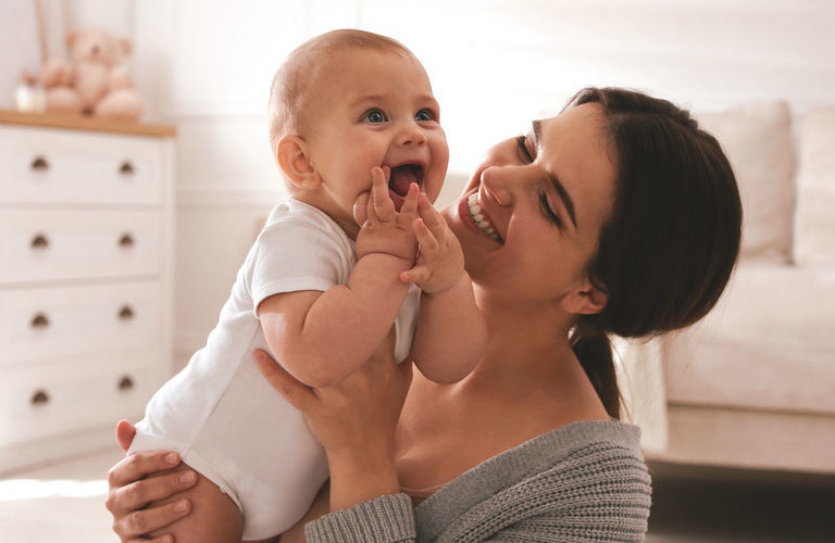 Mãe de primeira viagem de cabelos castanhos presos em um rabo de cavalo e sorridente, segurando no colo um bebê sorridente que veste body branco e coloca as mãos na boca.