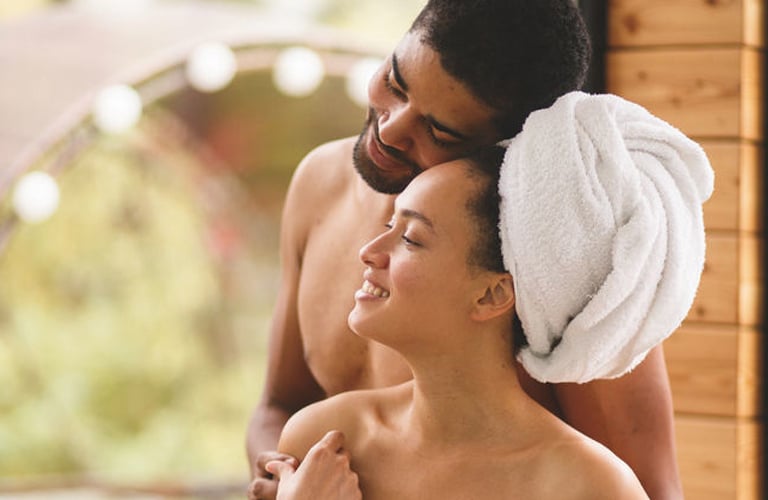 Homem e mulher abraçados e sorridentes em ambiente acolhedor de spa, vestindo toalhas brancas