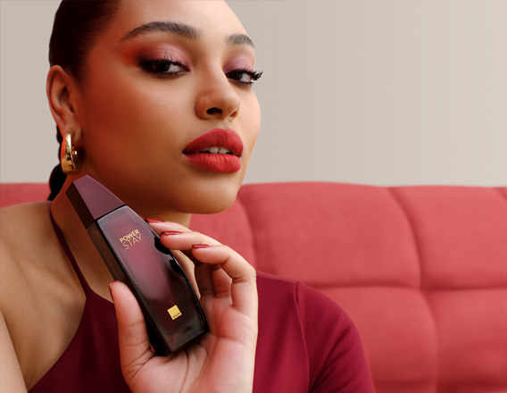 Mulher com maquiagem em tons de vermelho segura frasco de fragrância Power Stay em vermelho próximo ao  rosto. Ela usa vermelho e brincos dourados e está sentada em sofá vermelho em fundo claro.