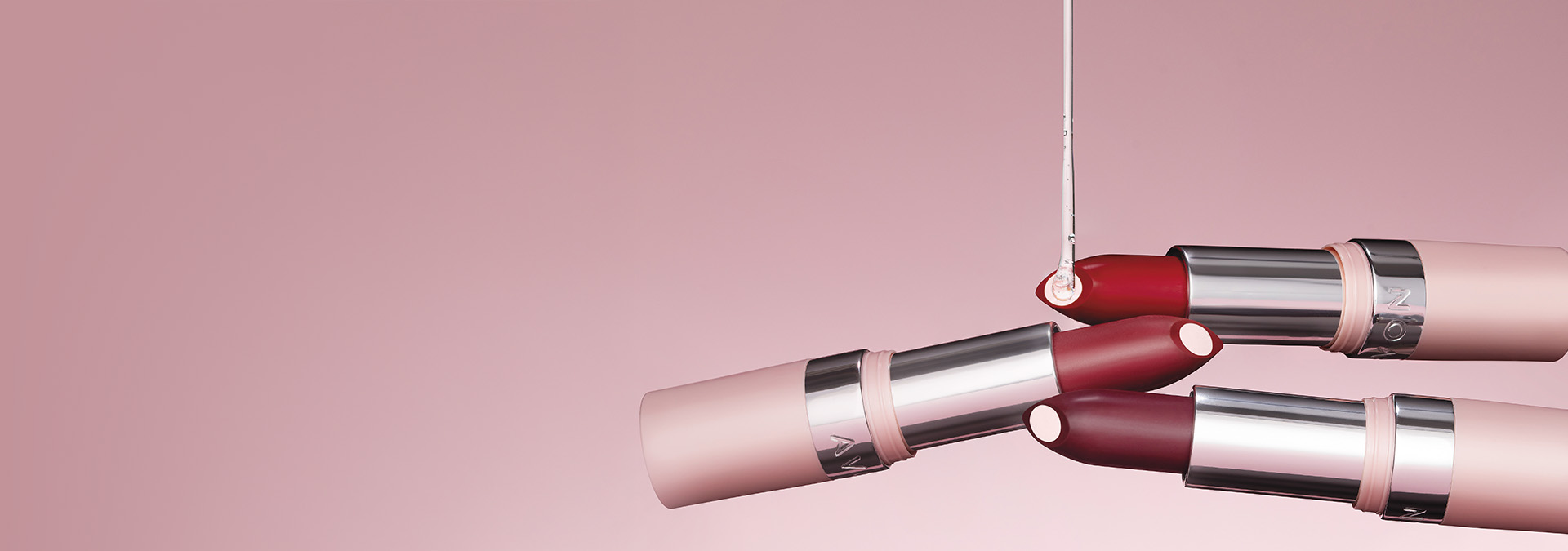 Banner com fundo rosa suave, apresentando batons em diferentes tons com embalagens elegantes. O texto destaca "Batom" e sugere hidratação e pigmentação intensa para um acabamento perfeito.