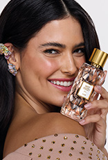 Mulher branca de cabelos castanhos segura frasco da fragrância Sweet Honesty Fun com tampa dourada e líquido translúcido. Ela sorri e está em fundo claro.
