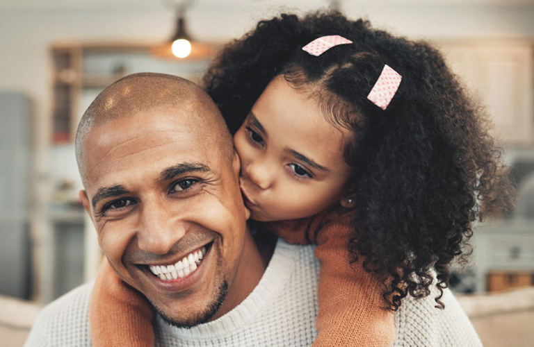 Um homem de cabeça raspada veste um suéter de tricô cinza-claro e sorri, enquanto sua filha está em suas costas, usando um blusão laranja e presilhas rosas no cabelo, enquanto o abraça. O fundo mostra o interior desfocado de uma casa aconchegante.