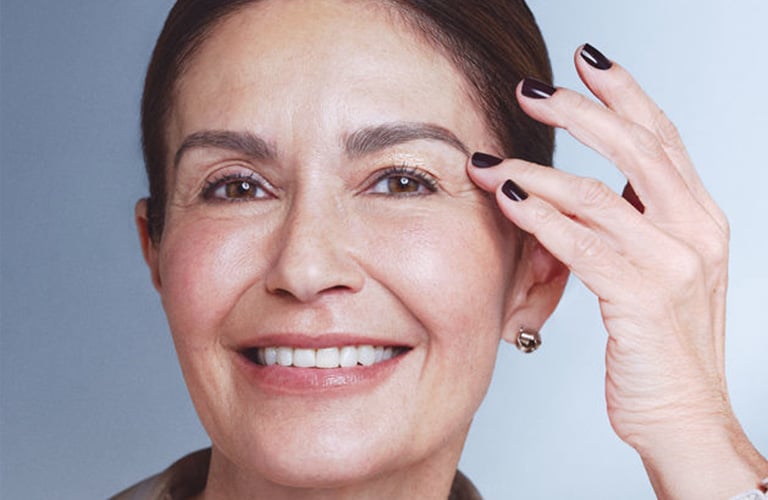 Mulher sorridente sênior, com cabelos castanhos presos em um coque e tocando a bolsa dos olhos com os dedos.