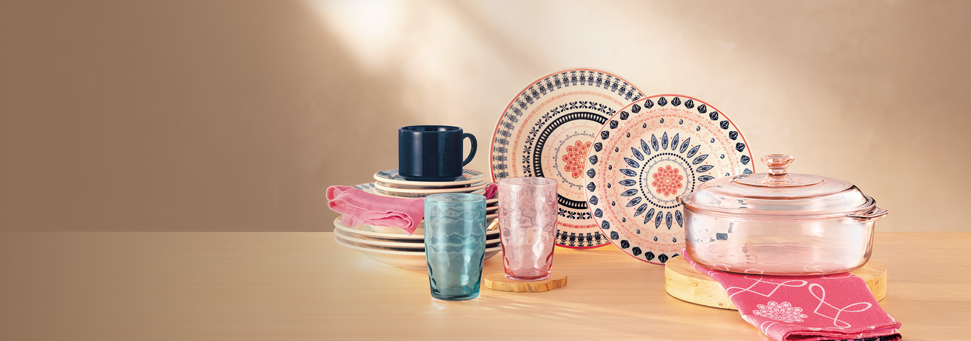 Banner com itens de cozinha sobre fundo neutro. Inclui pratos decorados, caneca azul, copos transparentes e uma panela de vidro. Texto: "Descubra a magia da cozinha com a gente!"