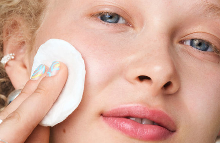 Mulher de cabelos loiros curtos está olhando diretamente para frente e limpa a bochecha com um algodão redondo branco.