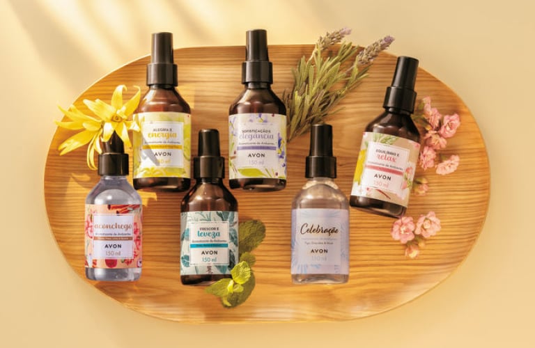 Kit de seis sprays aromatizadores da Avon em frascos escuros sobre bandeja de madeira para presente de casa nova.