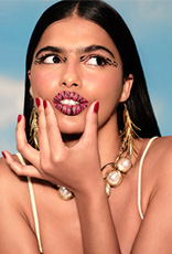 Mulher branca de cabelo preto escuro, usando delineado preto e arte animal print nos lábios. Ela pressiona as laterais da boca, olhando para o lado, em um fundo de céu azul claro.