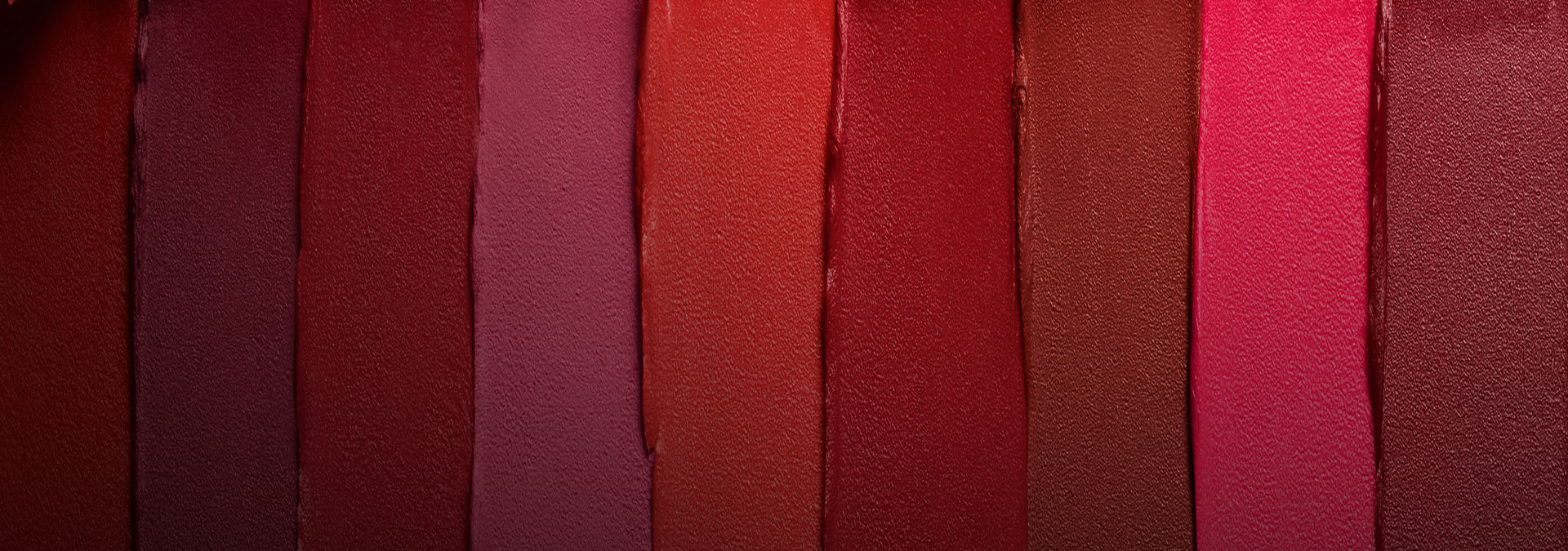 Banner com fundo em tons de vermelho e rosa. Elementos incluem faixas de batons aplicados em diferentes tonalidades, formando um degradê vibrante. Texto: "Maquiagem: mais presentes, mais emoção."