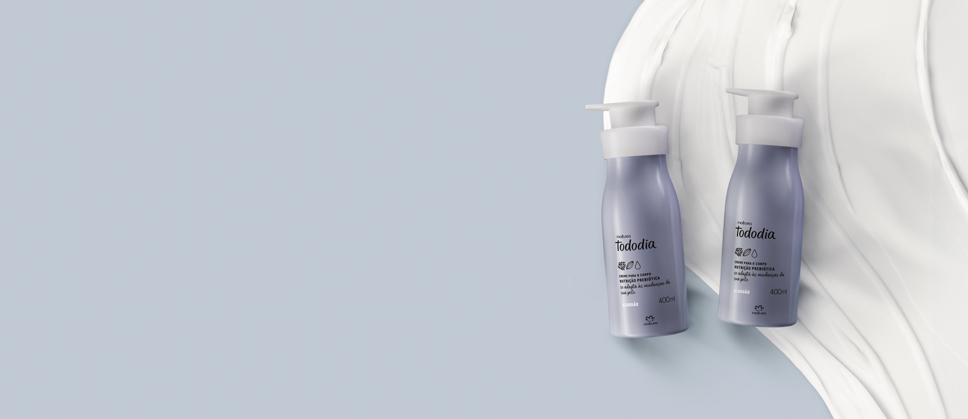 Dois frascos lilases com tampas pump brancas de Creme Nutritivo para o Corpo Tododia Algodão. Fundo azul suave com textura branca cremosa embaixo das embalagens.
