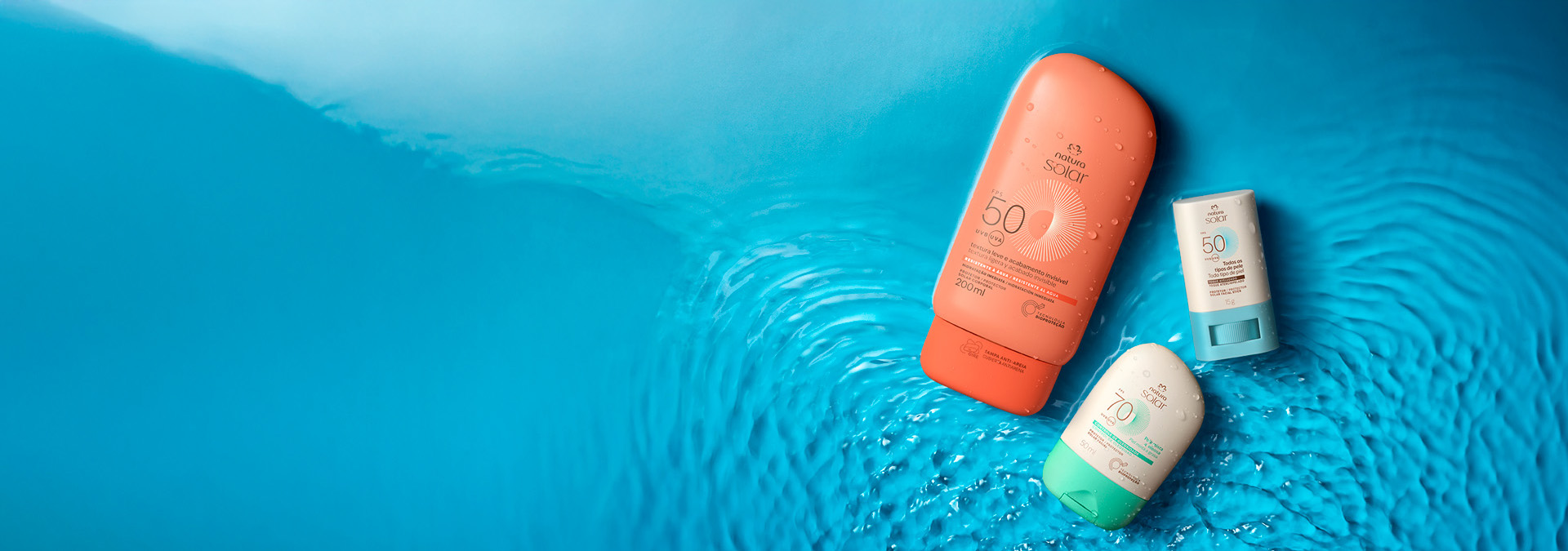 Três itens na diagonal em fundo azul aquoso: embalagem laranja de Protetor Solar Corporal, stick branco com azul de Protetor Solar Facial e embalagem branca com tampa verde de Protetor Solar Facial.
