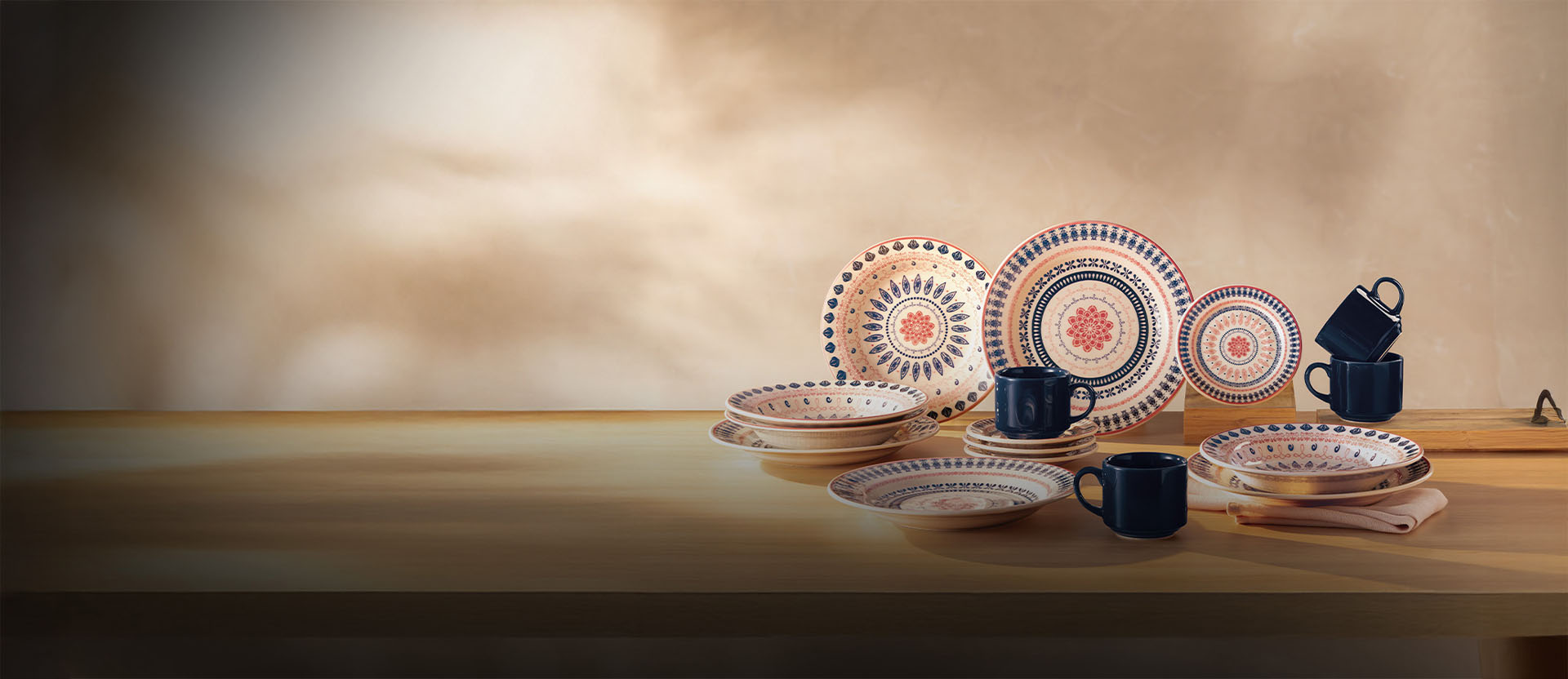 Banner com pratos e canecas sobre fundo neutro. As peças incluem pratos rasos e fundos com padrões geométricos em tons de azul e vermelho, além de canecas pretas. Texto: "Renove sua casa com estilo!"