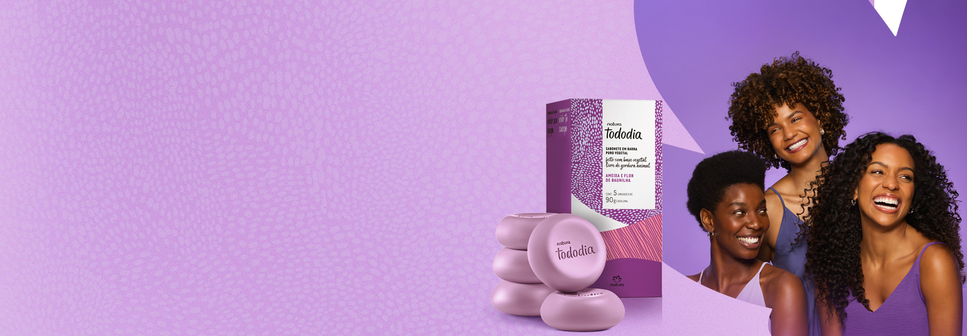 Duas mulheres negras, jovens e sorridentes, com cabelos encaracolados e crespos em fundo roxo. Ao lado, Sabonete em Barra Tododia Ameixa e Flor de Baunilha e cinco sabonetes redondos na cor roxa.