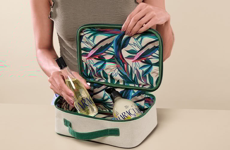 Foto de persona sobre fondo beige, abriendo un neceser rectangular con interior estampado de hojas y guardando frascos de productos líquidos en su interior.