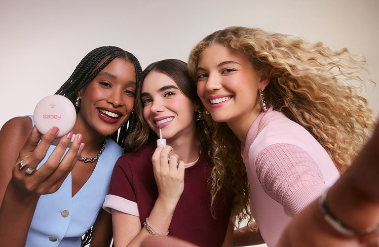 Tres amigas sonríen sobre un fondo claro. Una sostiene un producto de Natura Faces mientras otra se maquilla los labios.