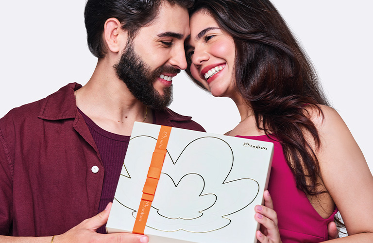 Foto sobre fondo blanco de una pareja abrazada sosteniendo una caja de regalo para mujer blanca con cinta naranja.