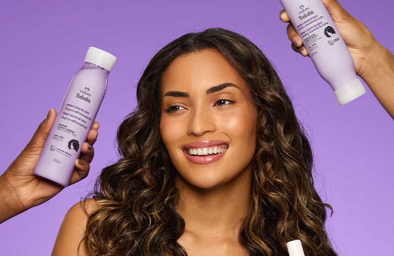 Foto de una persona de pie con el cabello rizado. Fondo lila uniforme. Alrededor, manos sostienen cuatro frascos lilas de productos para el cabello. La persona viste top lila y pantalón morado.