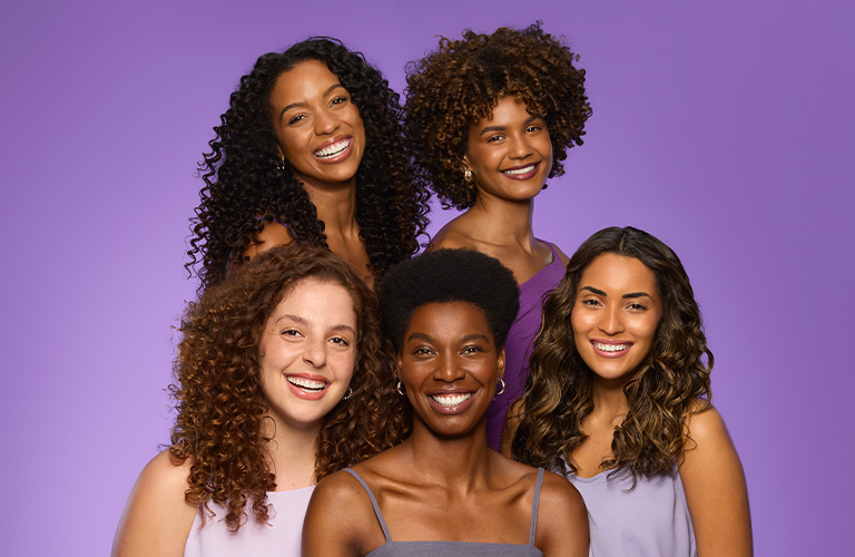 Foto de cinco mujeres sonriendo con distintos tipos de rulos y rizos, en dos filas. Fondo violeta.