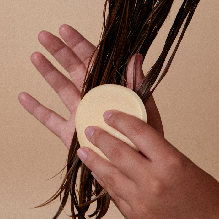 Una persona con cabello castaño lacio aplica un acondicionador en barra en el cabello. El fondo es beige claro.