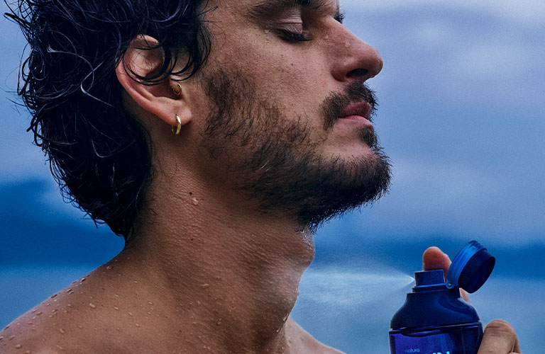 Un hombre sin camisa, con los ojos cerrados, rocía un perfume azul de la marca Kaiak. Fondo azul claro.