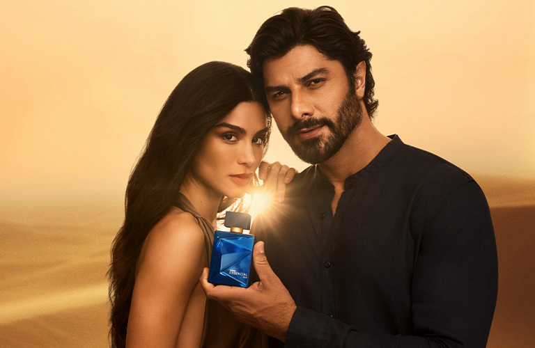 Foto con fondo dorado de desierto. Hombre y mujer juntos, mientras él sostiene un frasco azul del perfume Essencial Oud iluminado.
