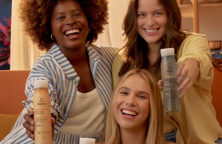 Foto de tres personas sentadas en un sillón. Fondo interior con cortina clara y estante. Elementos: frascos de productos para el cabello en las manos, en tonos rosa, beige y transparente.