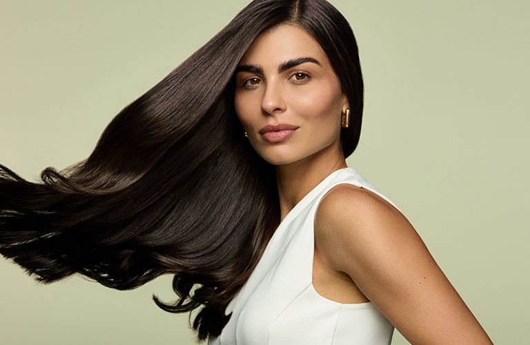 Foto de una mujer con cabello lacio y oscuro al viento. El fondo es verde claro. Lleva una blusa blanca sin mangas y usa aros dorados.