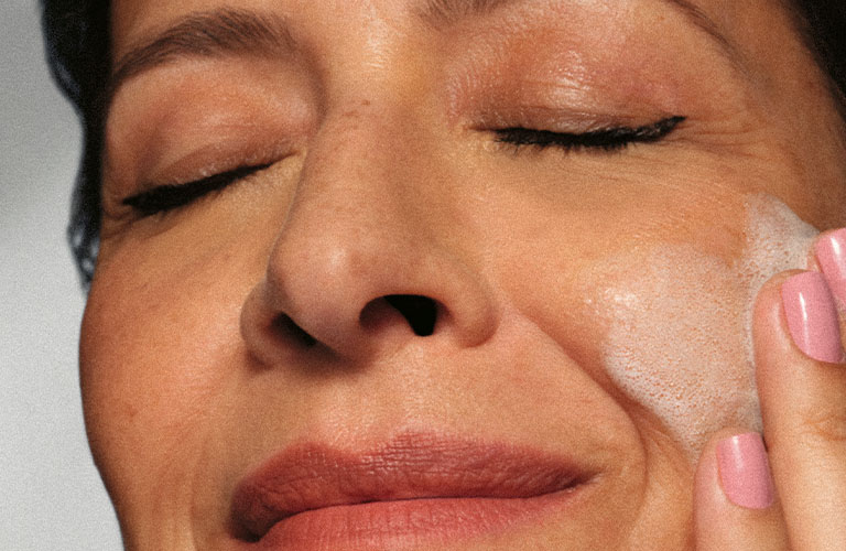 foto con fondo claro de una mujer con ojos cerrados aplicando crema en el rostro con la mano.