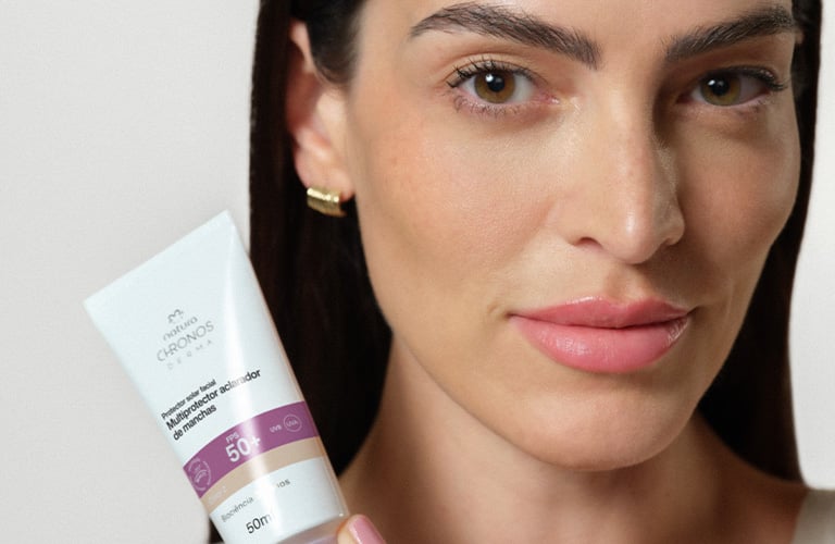 Foto con fondo claro. Persona sostiene un tubo de protector facial Chronos Derma FPS 50 frente al pecho; se ven mano, aro en la oreja y parte de un top claro.