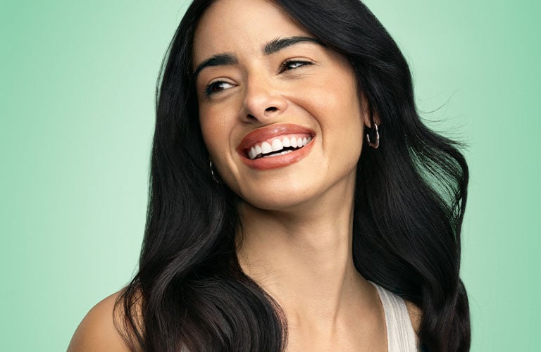 Foto de una mujer sonriendo, con un fondo verde liso. Lleva un top blanco y tiene el pelo largo y suelto sobre los hombros.