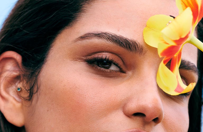 Foto con fondo celeste. Primer plano del lateral del rostro de una mujer, cabello oscuro, oreja con piercing y una flor naranja detrás de la oreja.