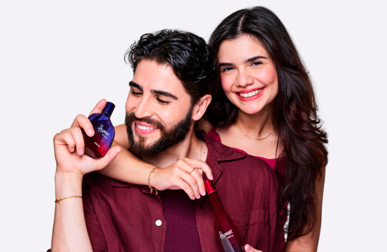 ) Foto con fondo gris claro. Un hombre y una mujer sonríen mientras muestran un frasco de perfume Natura Humor.