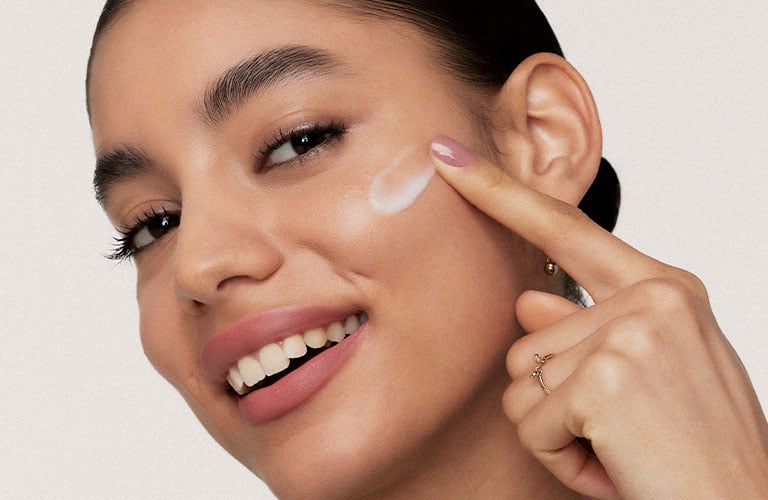 Foto con fondo claro. Una mujer con el cabello recogido lleva una camiseta verde con estampado blanco y señala con un dedo hacia su oreja.